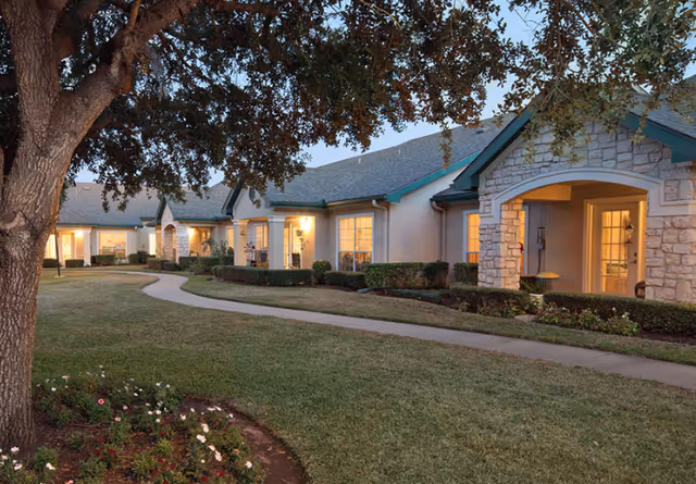 Exterior view of Carriage Inn Lake Jackson facility at dusk, showing a single-story building with stone and stucco facade, lit windows, a curved sidewalk, manicured lawn, flower beds, and a large tree in the foreground.