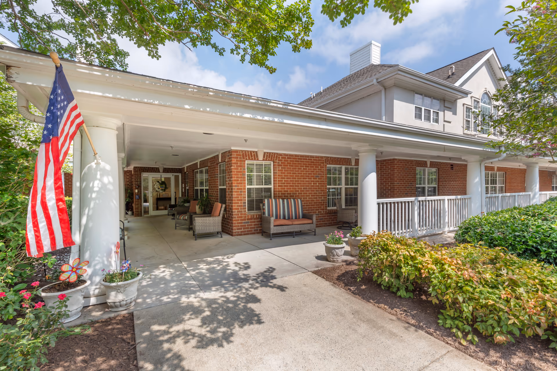 Covered front porch entrance of a brick senior living building with columns, seating, an American flag, and surrounding landscaping.