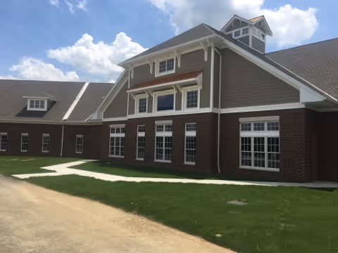 Exterior view of a senior living facility building with large windows, brick lower walls, and gray siding upper walls under a partly cloudy sky.