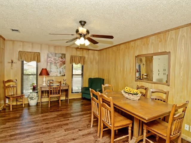 Wood-paneled dining room with a wooden table and six chairs, ceiling fan, small desk by the windows, and a green armchair.