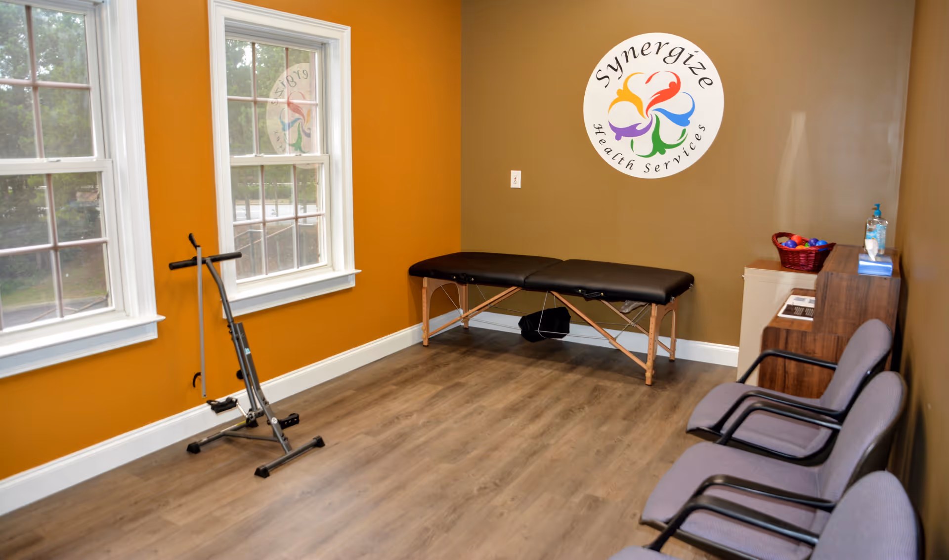 A therapy or treatment room with a black padded treatment table against a brown wall featuring a colorful Synergize Health Services logo. The room has two large windows with white trim, a piece of exercise equipment near the windows, three gray chairs with black armrests lined up along the right wall, and a small cabinet with a basket of colorful balls and hand sanitizer on top.