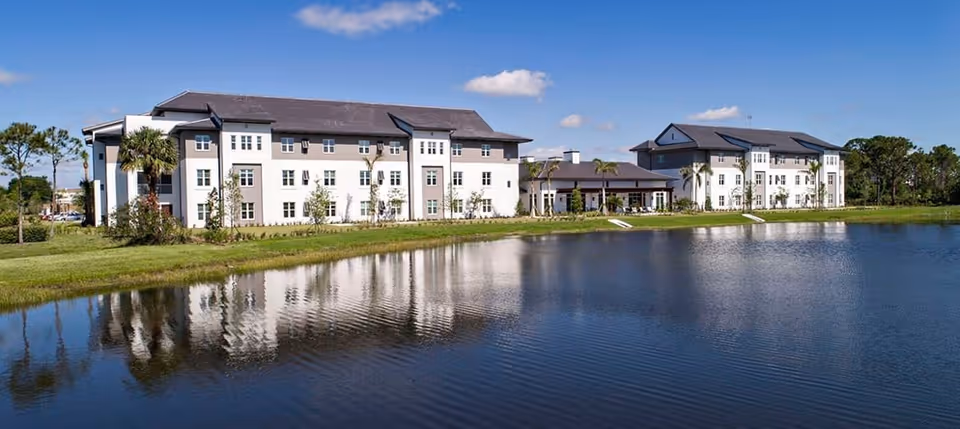A three-story senior living building and its reflection across a calm pond under a blue sky.