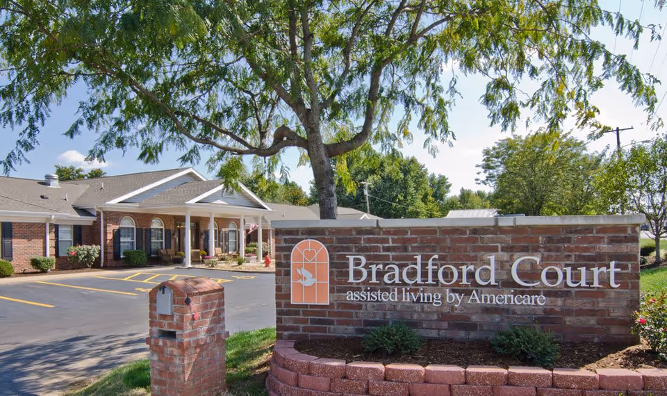 Exterior view of Bradford Court assisted living facility showing a brick sign with the facility name and a single-story building with a covered entrance, surrounded by trees and a parking area.