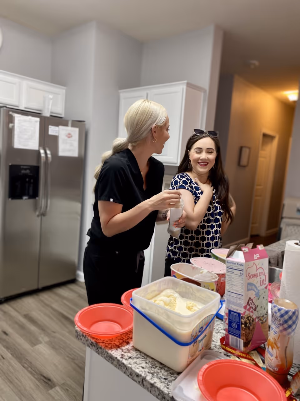 Two women standing in a kitchen, smiling and interacting near a counter with various food items including ice cream, bowls, and toppings. The kitchen has white cabinets and a stainless steel refrigerator.