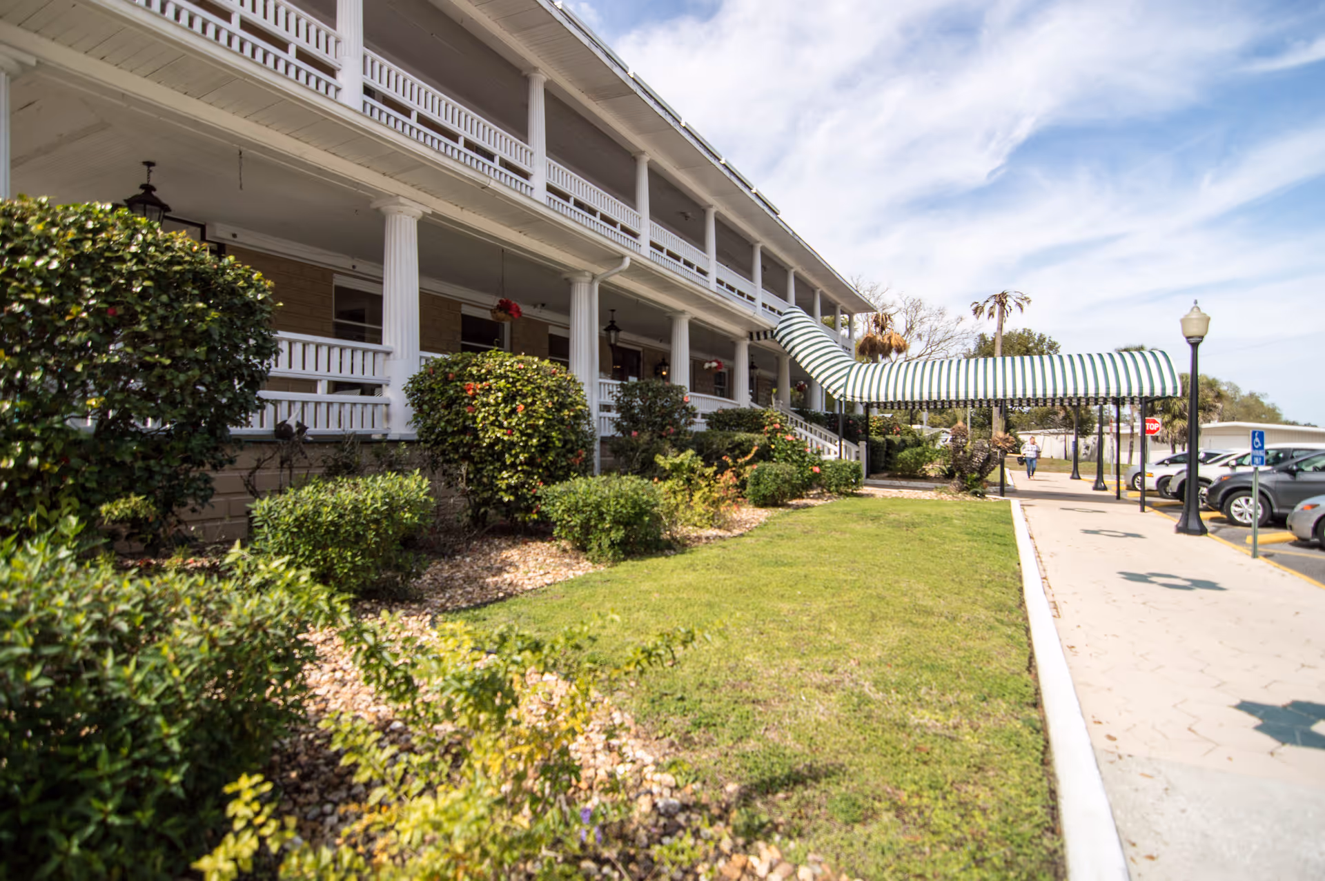 Exterior view of a senior living facility with a two-story white building featuring columns and a covered entrance with a striped awning. There are bushes and a grassy area in front, a sidewalk, and a parking lot with cars on the right side under a partly cloudy sky.