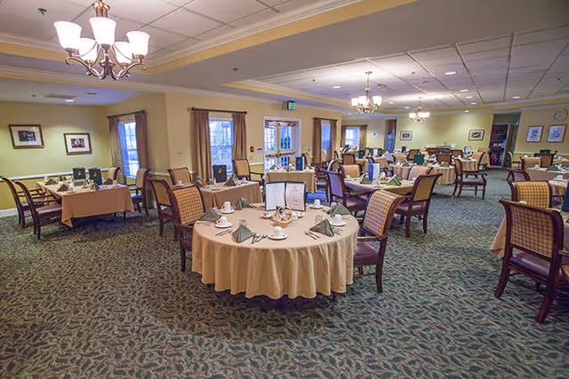 A spacious dining room with multiple round and rectangular tables set with beige tablecloths, green folded napkins, white cups and saucers, and menus. The room has patterned carpet flooring, beige walls, several windows with curtains, and chandeliers hanging from the ceiling providing warm lighting.