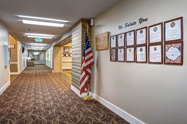 A long, carpeted hallway in a senior living facility with beige walls and ceiling lights. On the right wall, there is a display titled 'We Salute You' featuring framed certificates and plaques honoring veterans, along with an American flag on a stand. The hallway extends into the distance with doors and seating areas visible.