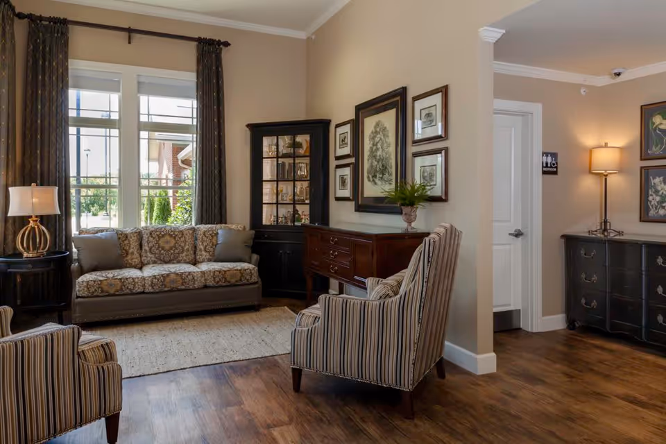 A cozy living room area with a patterned sofa, two striped armchairs, a wooden cabinet with framed artwork above it, a glass display cabinet, and a side table with a lamp. The room has large windows with dark curtains, wood flooring, and a doorway leading to a restroom.