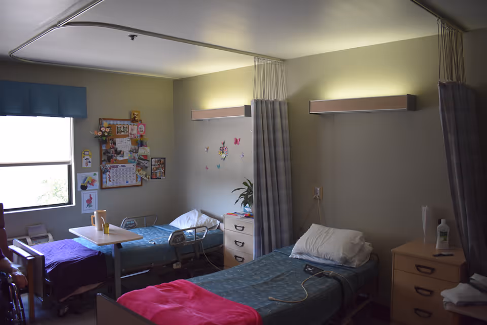 A shared hospital-style room with two beds separated by a curtain. Each bed has a bedside table with drawers and a wall-mounted light above. One bed has a pink blanket, the other has a purple blanket. There is a window with a blue valance, a bulletin board with photos and notes on the wall, and a small table with a pitcher and cup between the beds.