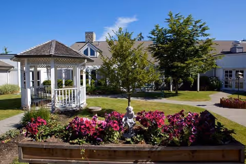 Outdoor garden area at Garden Courte Memory Care featuring a white gazebo, a flower bed with vibrant pink flowers and a small statue, a tree, and a building in the background under a clear blue sky.