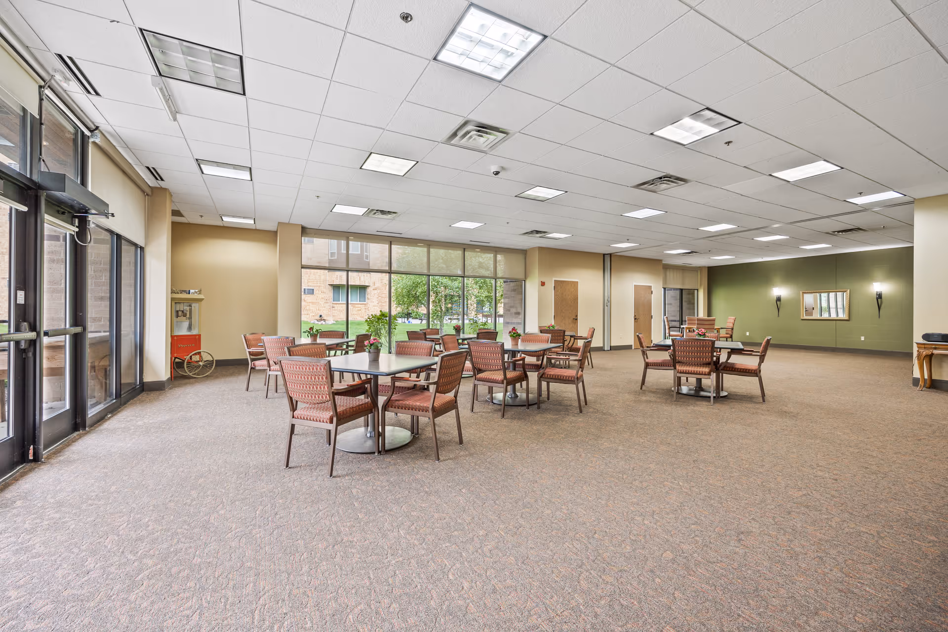 A spacious senior living facility common area with multiple tables and chairs arranged for seating. The room has large windows letting in natural light, beige walls, and a carpeted floor. There is a popcorn machine in the corner near the windows and a small table with a radio on the right side of the room.