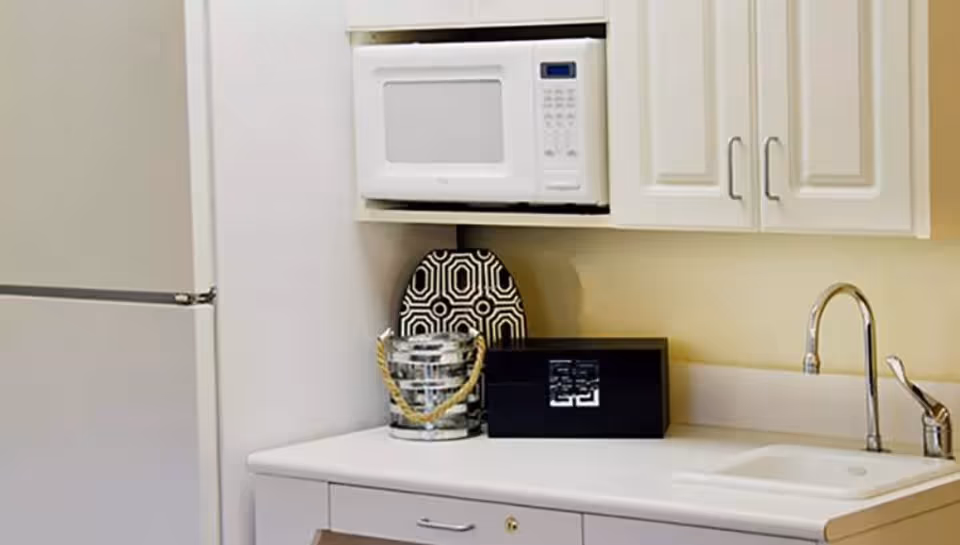 A small kitchen area featuring a white refrigerator, a white microwave mounted above the counter, a decorative black and white plate, a small silver bucket with rope handles, a black box, and a white sink with a chrome faucet.