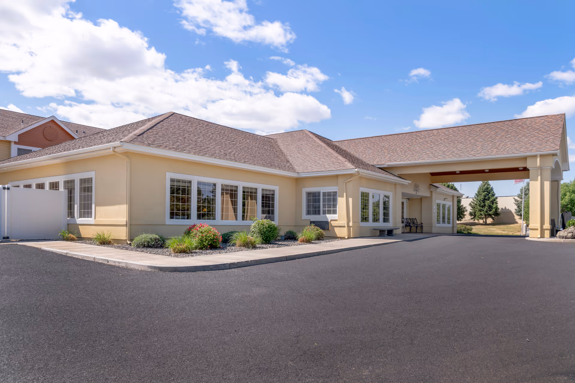 Exterior view of a single-story building with beige walls and a brown shingled roof under a partly cloudy blue sky. The building has multiple white-framed windows and a covered entrance with columns. There are small landscaped areas with bushes and flowers near the building, and a paved driveway in front.