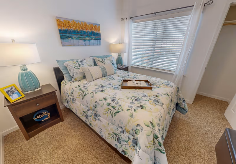 A cozy bedroom with a floral patterned bedspread on a double bed, two bedside tables each with a lamp, a window with white blinds and curtains, and a painting of trees above the bed.