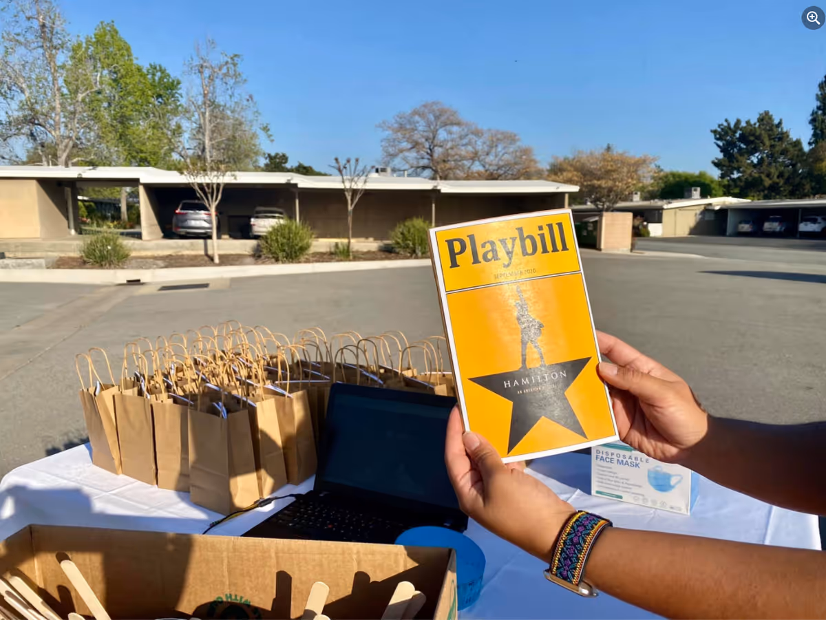A person holding a Playbill for the musical Hamilton outdoors near a table with brown paper gift bags, a laptop, and a box of disposable face masks. In the background, there are parked cars and single-story buildings under a clear blue sky.