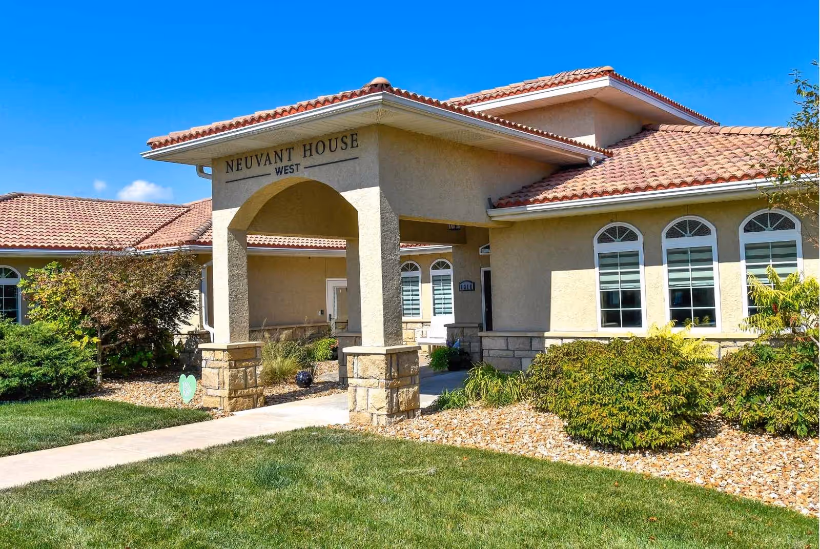 Exterior view of Neuvant House West, a single-story building with beige stucco walls and a red tile roof. The entrance features a covered walkway supported by stone pillars. The surrounding area includes green grass, bushes, and small trees under a clear blue sky.