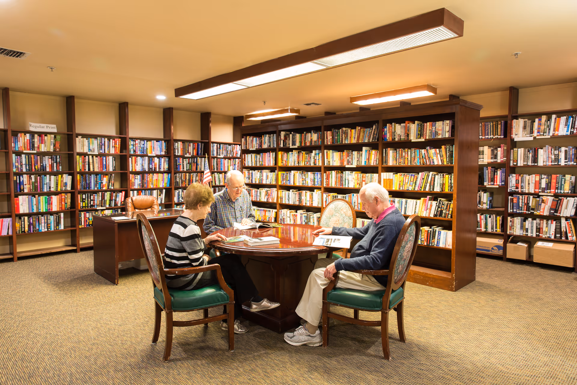Three elderly people sitting around a wooden table in a library room, reading books. The room has shelves filled with books along the walls and warm lighting from ceiling fixtures.