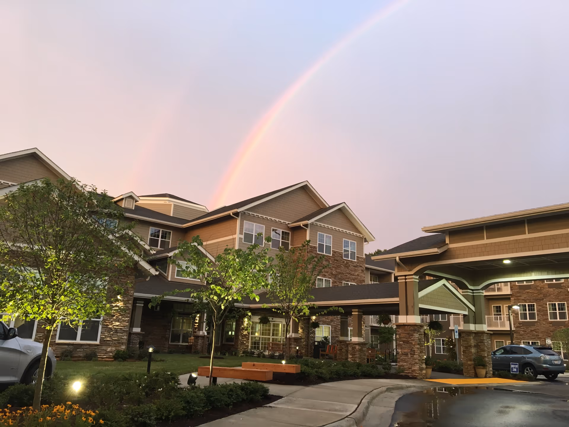 Exterior view of a multi-story senior living facility with stone and beige siding, surrounded by small trees and landscaping. A covered entrance with pillars is visible, and a double rainbow appears in the sky above the building.