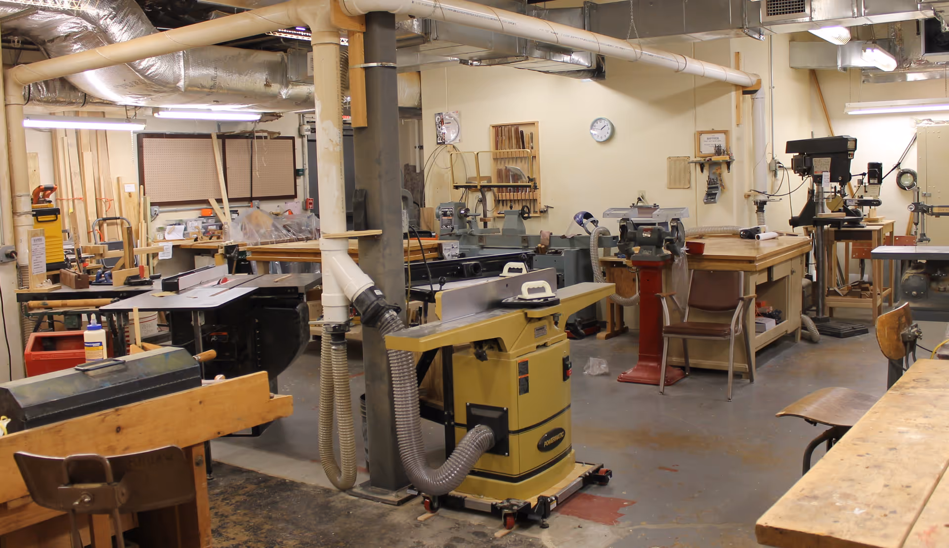 A woodworking workshop with various tools and machines including a table saw, drill press, and workbenches. The room has exposed pipes and ductwork on the ceiling, wooden chairs, and pegboards with tools hanging on the walls.