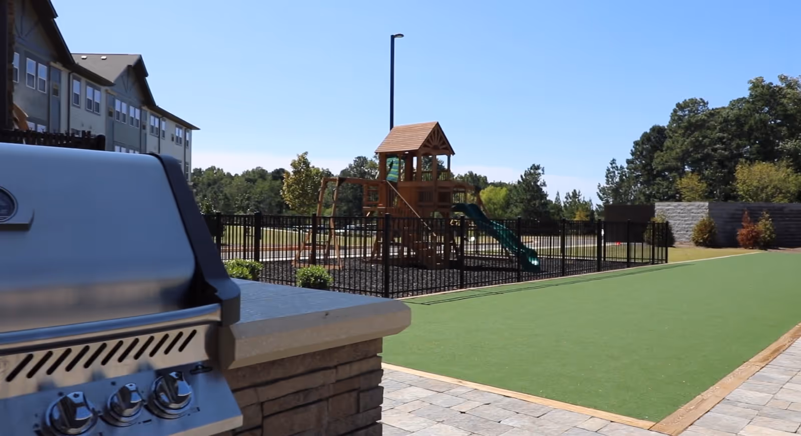 Outdoor area at The Phoenix at Union Hill featuring a stainless steel barbecue grill in the foreground, a fenced wooden playground with a slide in the background, and a green artificial turf area adjacent to a paved walkway under a clear blue sky.