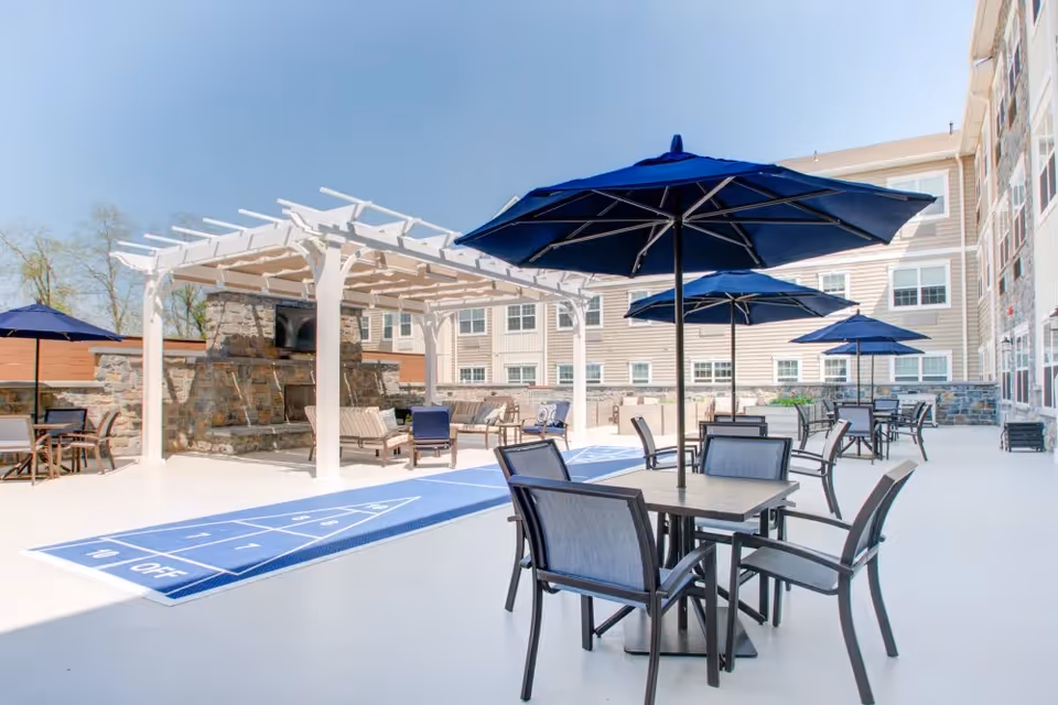 Outdoor patio area at Merrill Gardens at Glen Mills featuring multiple tables with chairs and blue umbrellas, a shuffleboard court, and a white pergola with seating and a stone fireplace in the background.