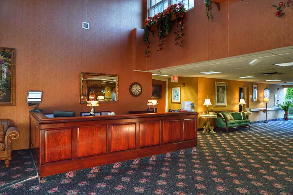 Reception area of a nursing and rehabilitation center with a large wooden front desk, a wall clock, a mirror, floral carpet, and seating area with green upholstered chairs and lamps. The walls are decorated with framed artwork and floral arrangements hang from a ledge near the ceiling.