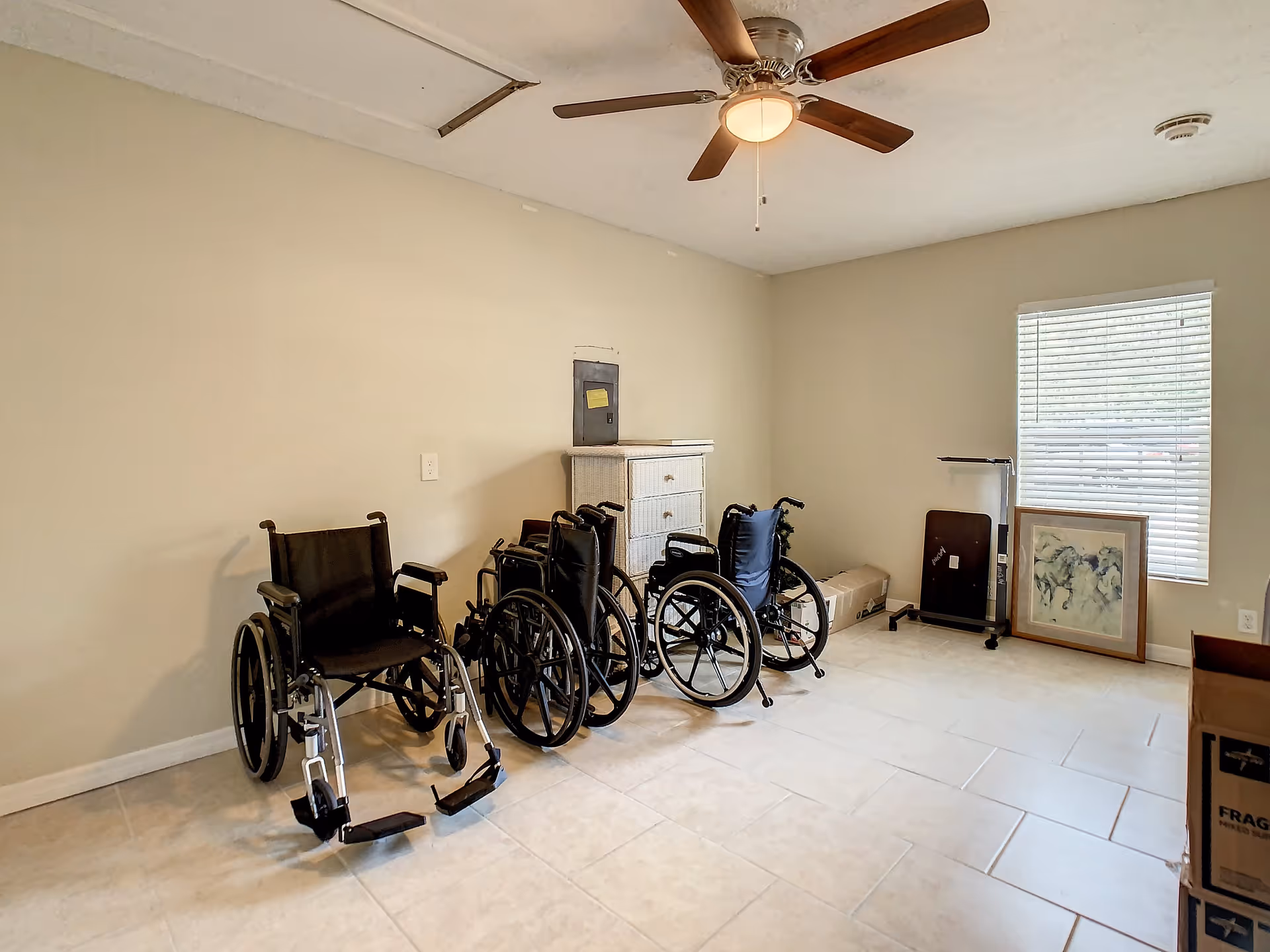 A room with beige walls and tiled floor containing four wheelchairs lined up against one wall, a white chest of drawers, a ceiling fan with light, a window with blinds, a framed picture resting on the floor, and some boxes.