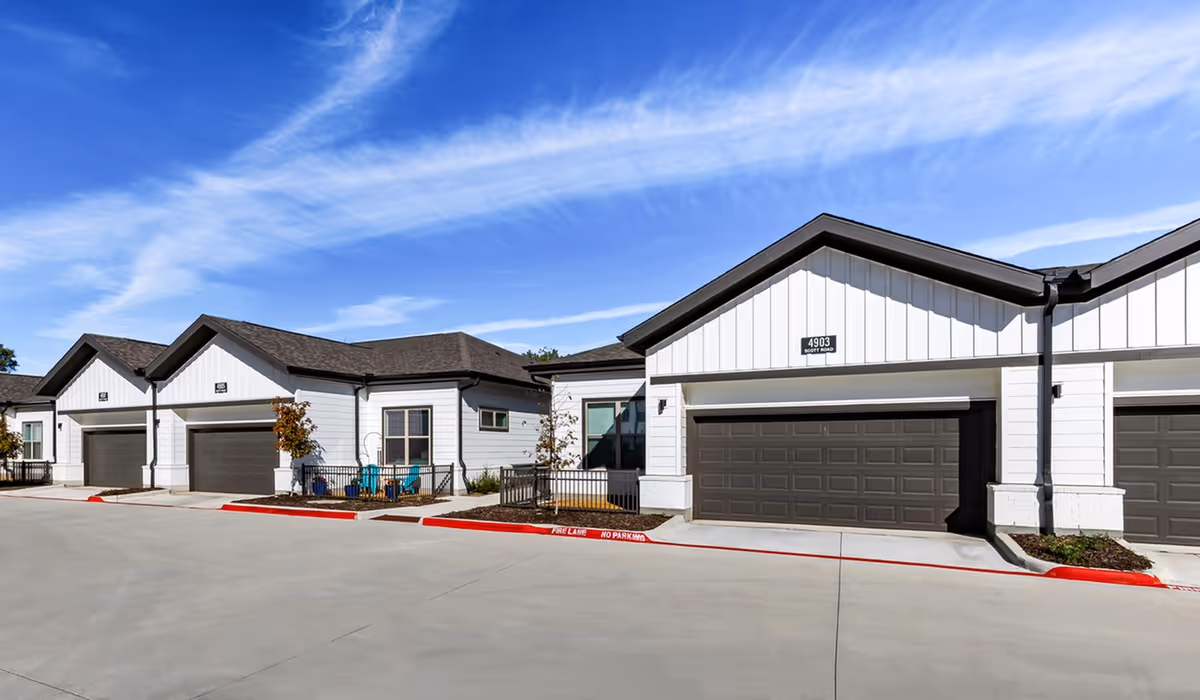 Exterior view of a modern senior living facility with white siding and dark brown garage doors under a bright blue sky with wispy clouds. The buildings have small fenced patios with outdoor chairs and young trees planted along the sidewalk.