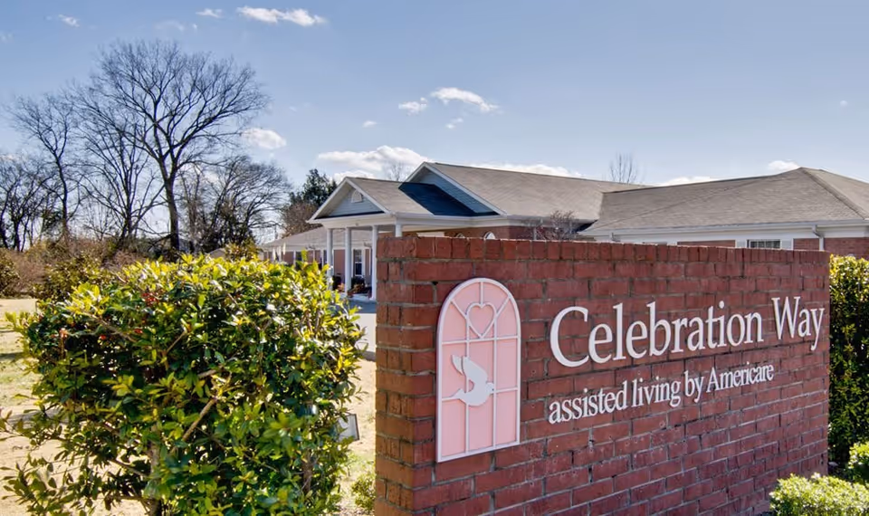 Exterior view of Celebration Way assisted living facility showing a brick sign with the facility name and logo, surrounded by greenery and trees under a clear blue sky.