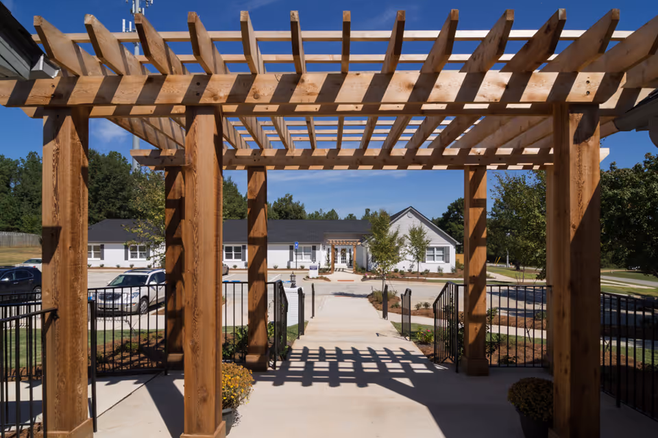 View through a wooden pergola structure looking out towards a white single-story building with a black roof, surrounded by trees and a parking lot with several cars under a clear blue sky.