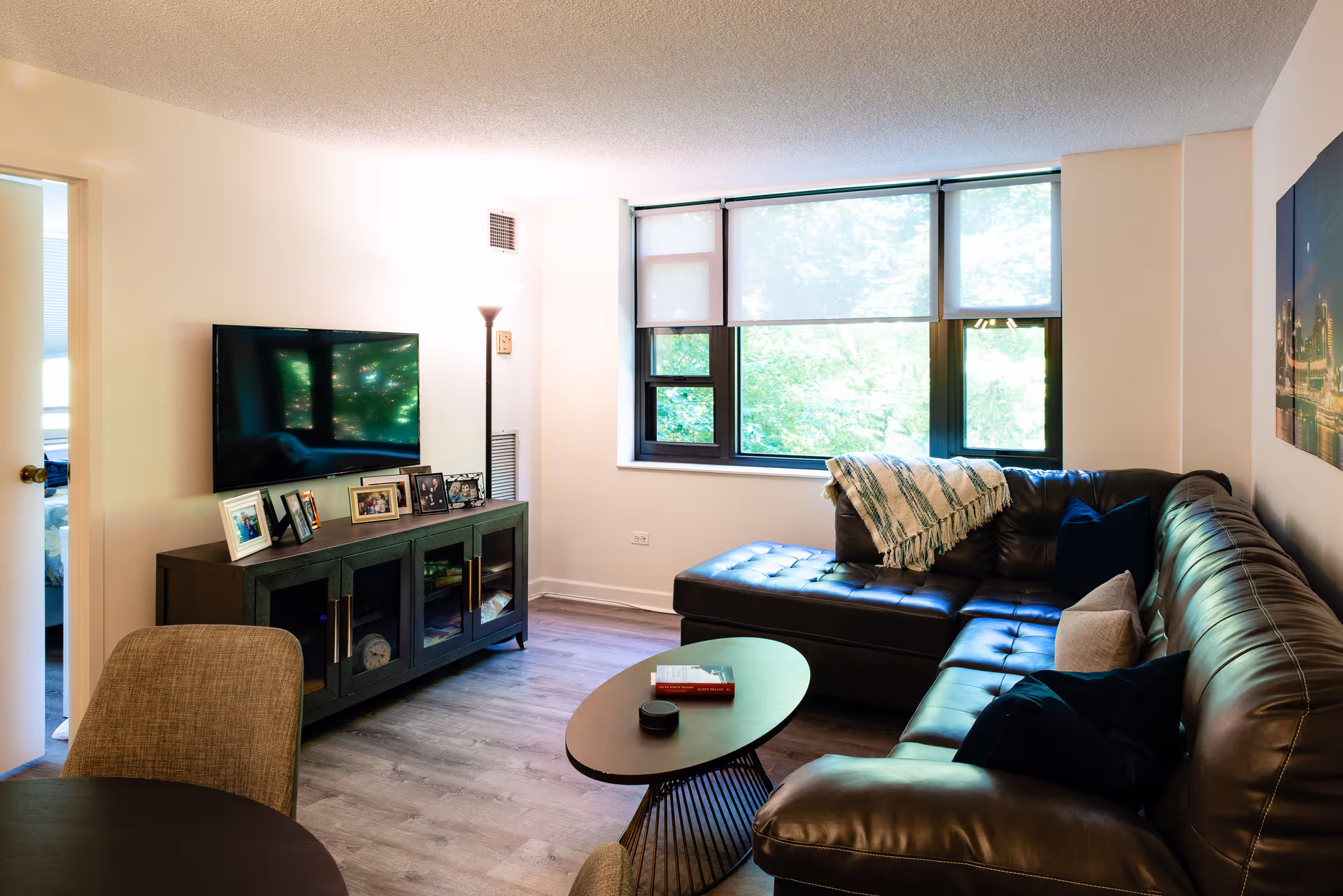 Bright living room with a dark leather sectional sofa, coffee table, TV on a media console, and a large window.