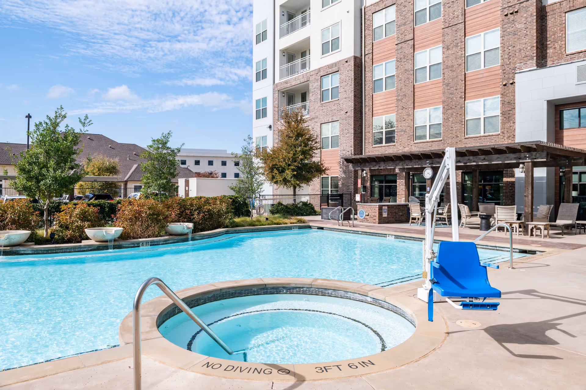 Outdoor swimming pool area at Overture Highlands with a round hot tub in the foreground, a blue pool lift chair, and a multi-story building with balconies and a shaded seating area in the background under a partly cloudy sky.