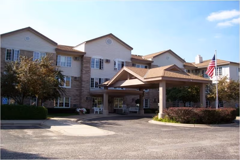 Exterior view of a multi-story senior living facility with a covered entrance, surrounded by trees and bushes, and an American flag flying on a flagpole under a partly cloudy sky.