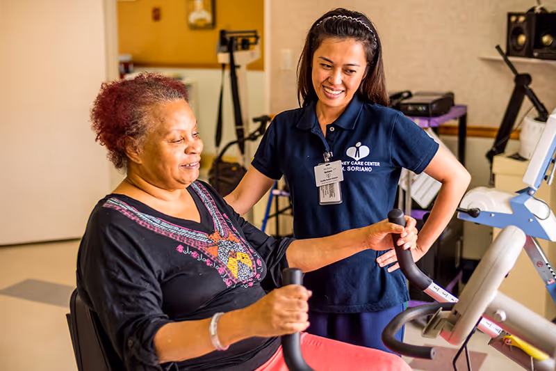 A senior woman is exercising on a stationary bike while a female caregiver or physical therapist stands beside her, smiling and providing support in a care facility room.