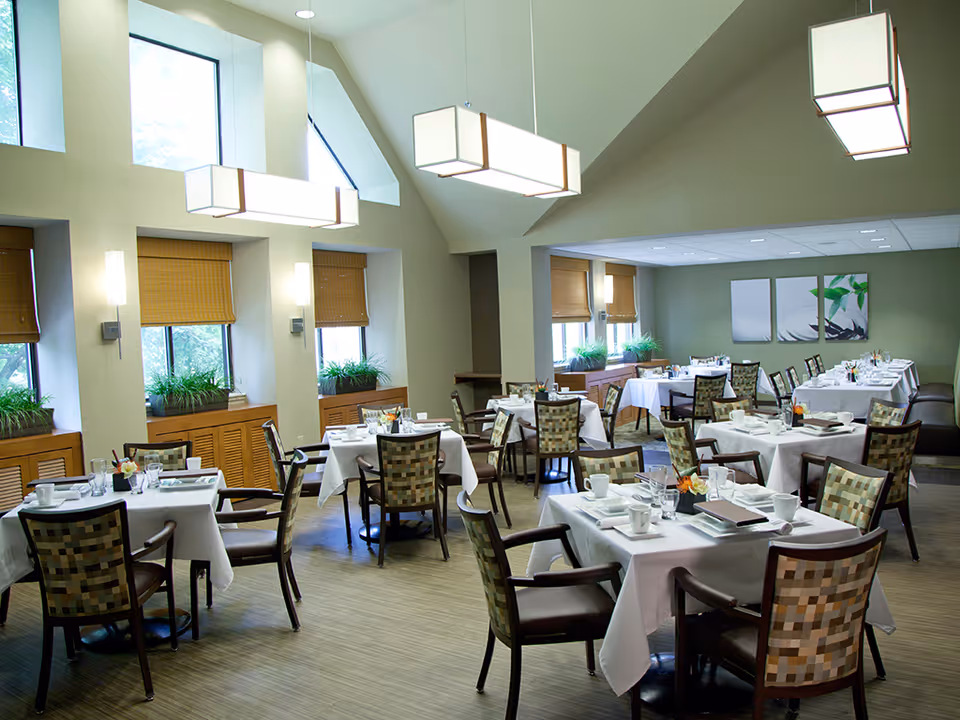 A bright and spacious dining room with multiple tables covered in white tablecloths, each set with cups, glasses, and napkins. The room features large windows with bamboo blinds, green plants on the window sills, modern pendant lights hanging from a high ceiling, and patterned cushioned chairs around the tables. There are three framed artworks on the far wall and a bench seating area along one side of the room.
