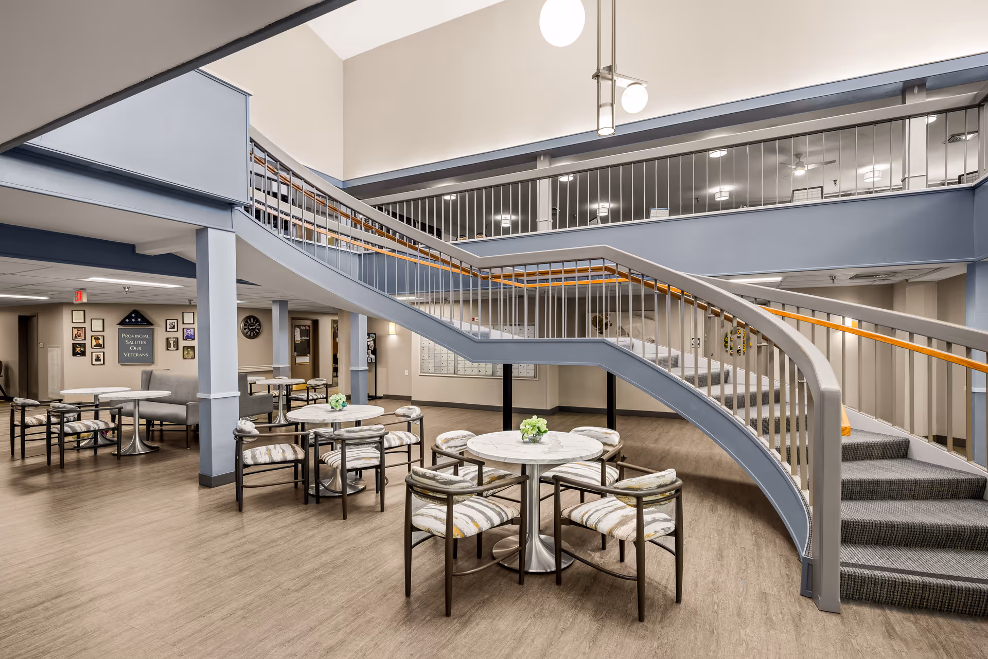 Interior view of a senior living community common area featuring a curved staircase with gray railings and wooden handrails. The space includes several round tables with cushioned chairs arranged around them. The walls are decorated with framed pictures and a sign that reads 'Provincial Salutes Our Veterans.' The flooring is a light wood tone, and the area is well-lit with ceiling lights.