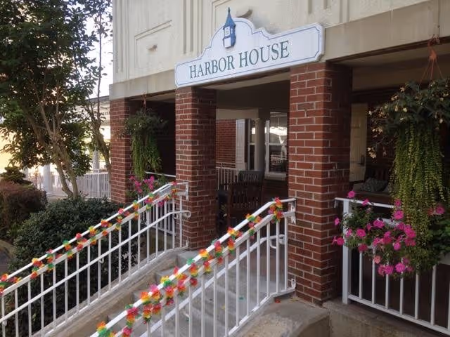Entrance of Harbor House assisted living showing a ramp with railings, brick columns, hanging plants, and a sign above the doorway.