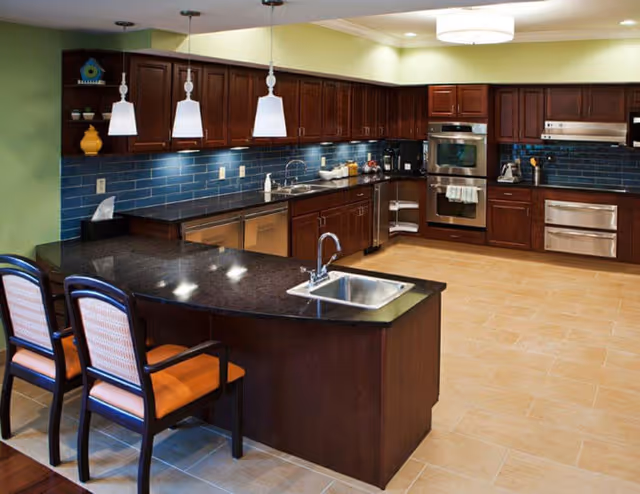 A modern kitchen with dark wood cabinets, a black countertop island with a built-in sink, and three pendant lights hanging above. The backsplash is made of blue tiles, and the kitchen features stainless steel appliances including an oven, microwave, and dishwasher. Two chairs with orange cushions are placed at the island.