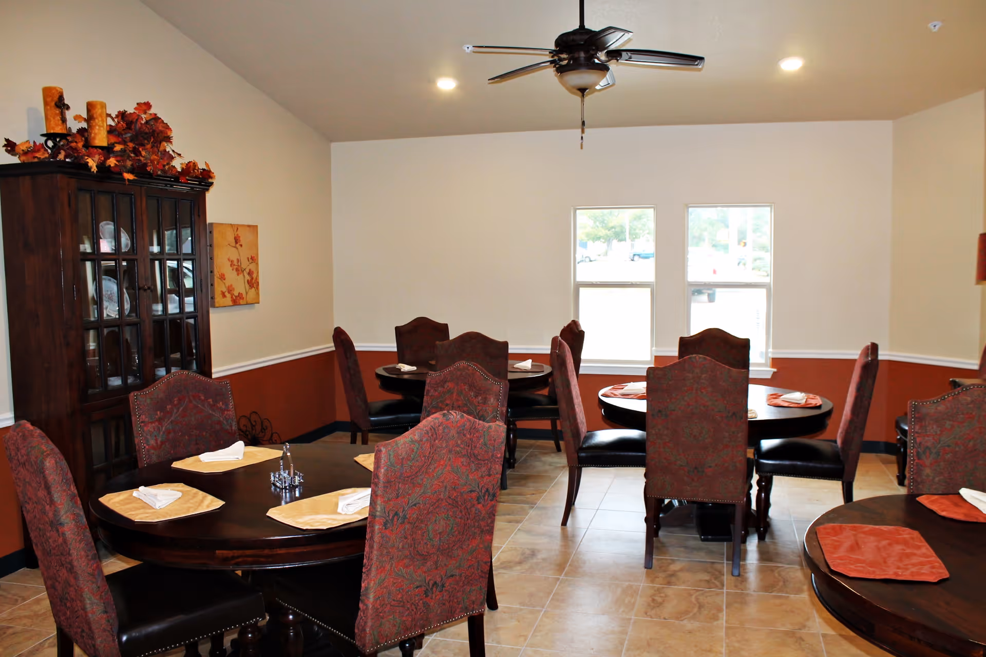 Dining room with multiple round wooden tables, each set with placemats and folded napkins. The chairs have patterned upholstery and dark wooden legs. There is a dark wooden cabinet with glass doors on the left side, decorated with candles and autumn leaves on top. The walls are painted beige with a dark red lower section and two windows letting in natural light.