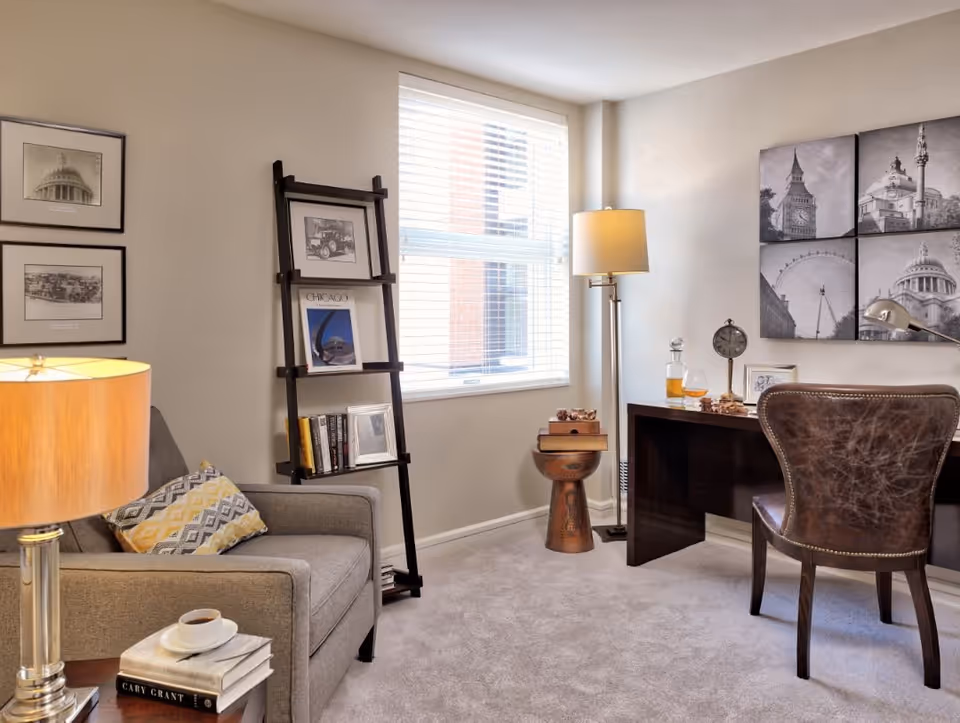 Cozy living room/office featuring an armchair with a side table, a leaning ladder bookshelf, a desk with a leather chair, and a window letting in natural light.