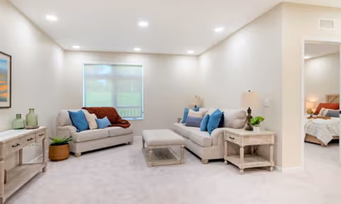 A bright living room with two beige sofas adorned with blue and beige pillows, a matching ottoman in the center, two side tables with lamps and plants, a console table with decorative glass bottles, and a window with a view of greenery. An adjacent bedroom is partially visible through an open doorway.