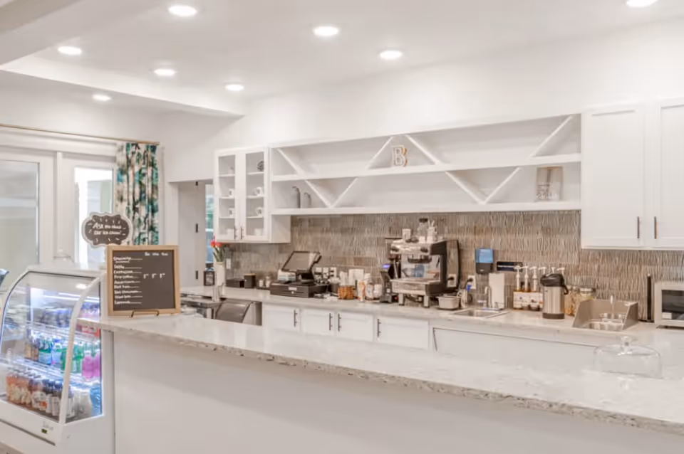 Bright and clean cafe counter area with a glass display case containing beverages and snacks, a menu board, coffee machine, and various coffee supplies on the countertop. White cabinets and shelves are mounted on the wall behind the counter, with a tiled backsplash and recessed ceiling lights illuminating the space.