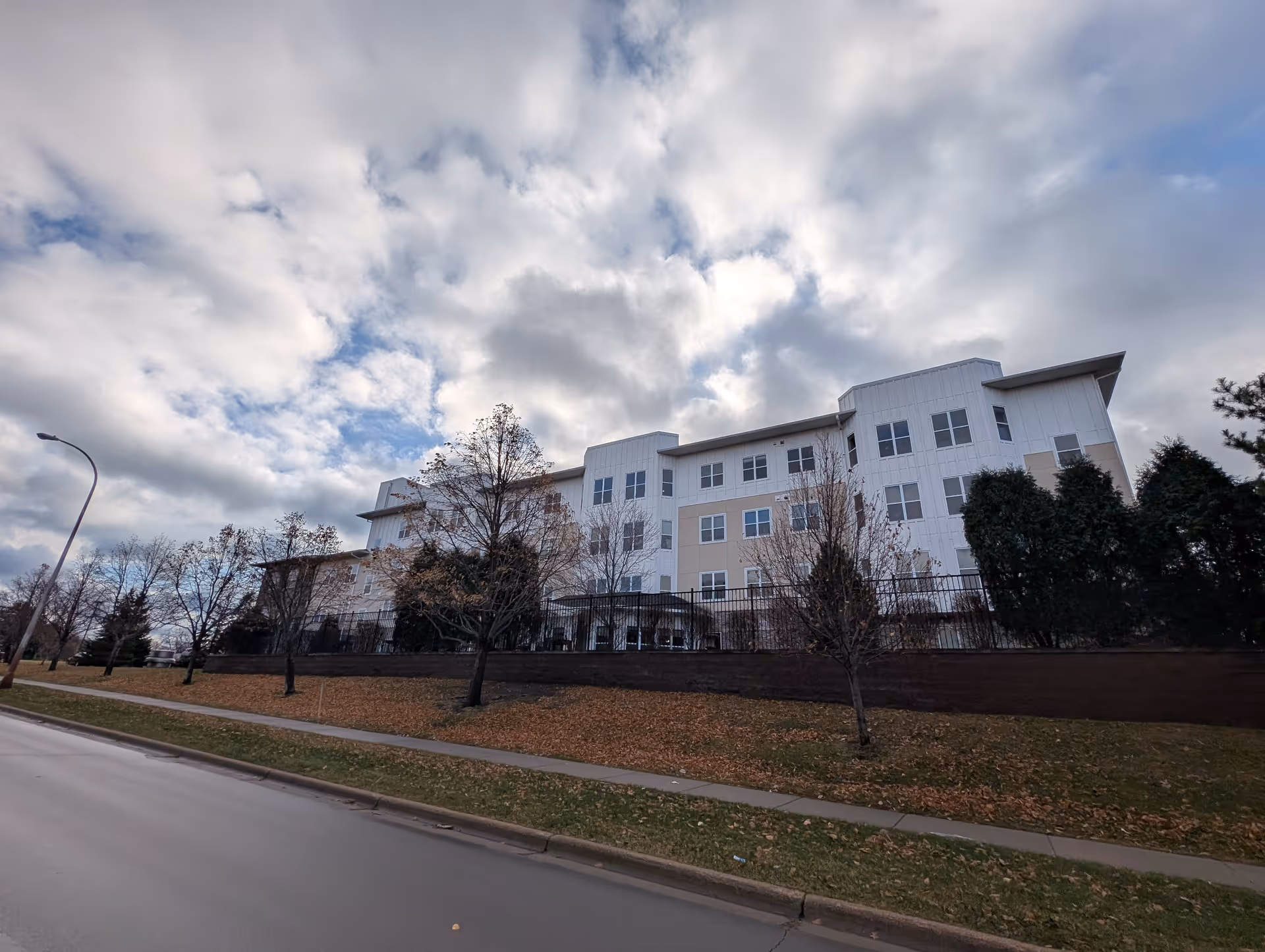Exterior view of a multi-story senior living facility building with beige and white siding, several windows, and a fenced area in front. Leafless trees and some evergreen shrubs are visible along the sidewalk and grassy area under a partly cloudy sky.