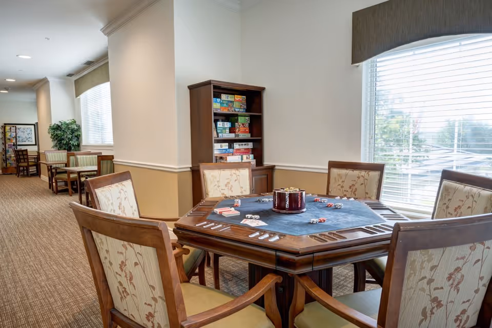 A well-lit common area with a hexagonal wooden game table in the foreground, surrounded by six cushioned chairs with floral upholstery. The table has poker chips and playing cards on it. In the background, there are additional tables and chairs along a carpeted hallway, a bookshelf filled with board games, a large window with blinds, and a potted plant.