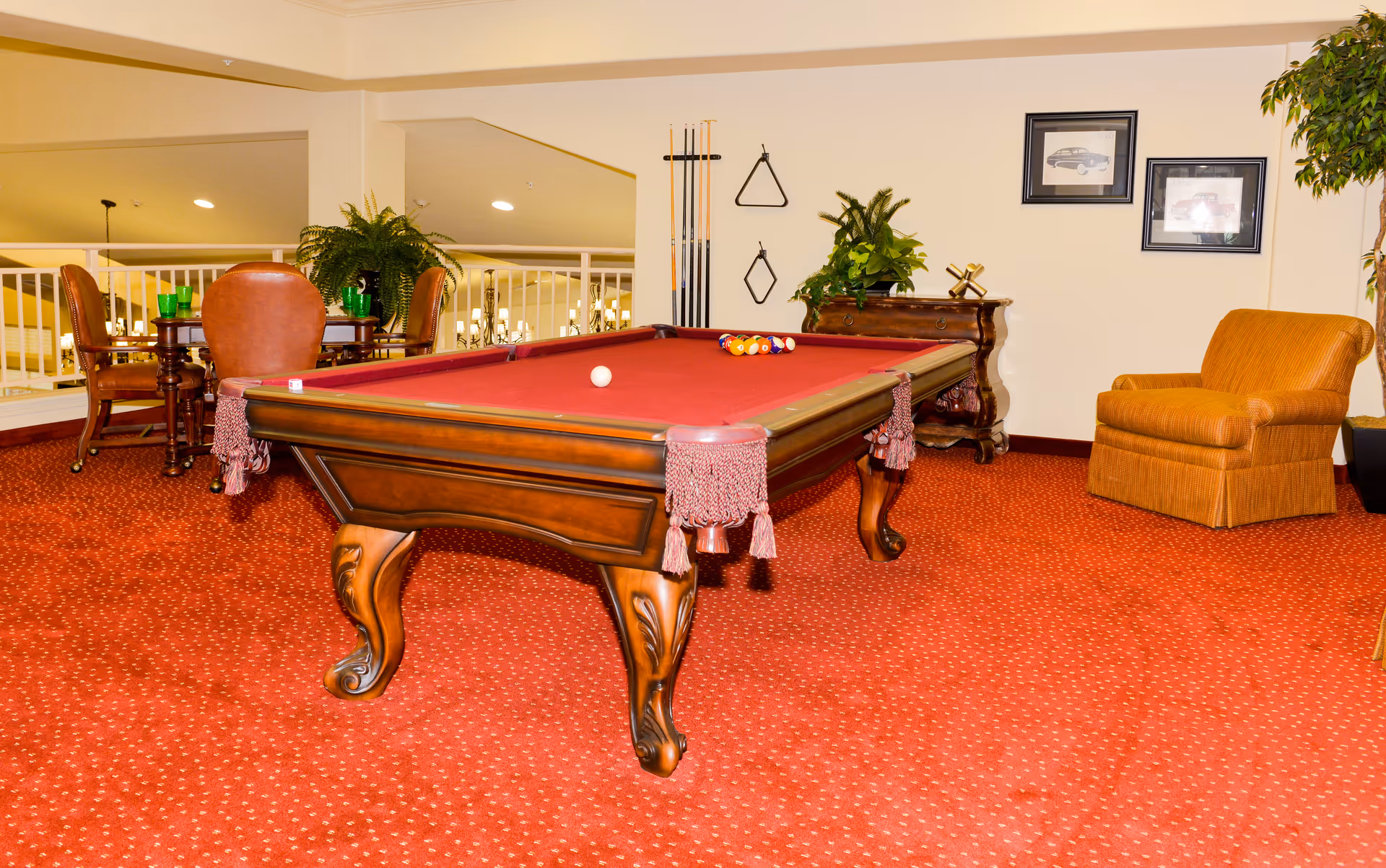 A cozy indoor recreational area featuring a red felt pool table with balls and a cue ball arranged for a game. The room has red patterned carpet, a wooden side table with plants and decorative items, a comfortable orange armchair, and a small dining table with chairs in the background. Pool cues and racks are mounted on the wall, along with framed pictures and additional plants.