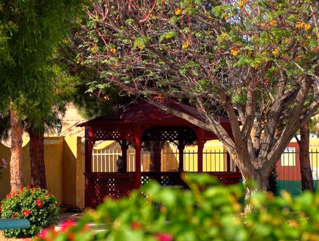 A red wooden gazebo surrounded by trees and green bushes with some flowers in an outdoor garden area. The background shows a yellow wall and a metal fence.