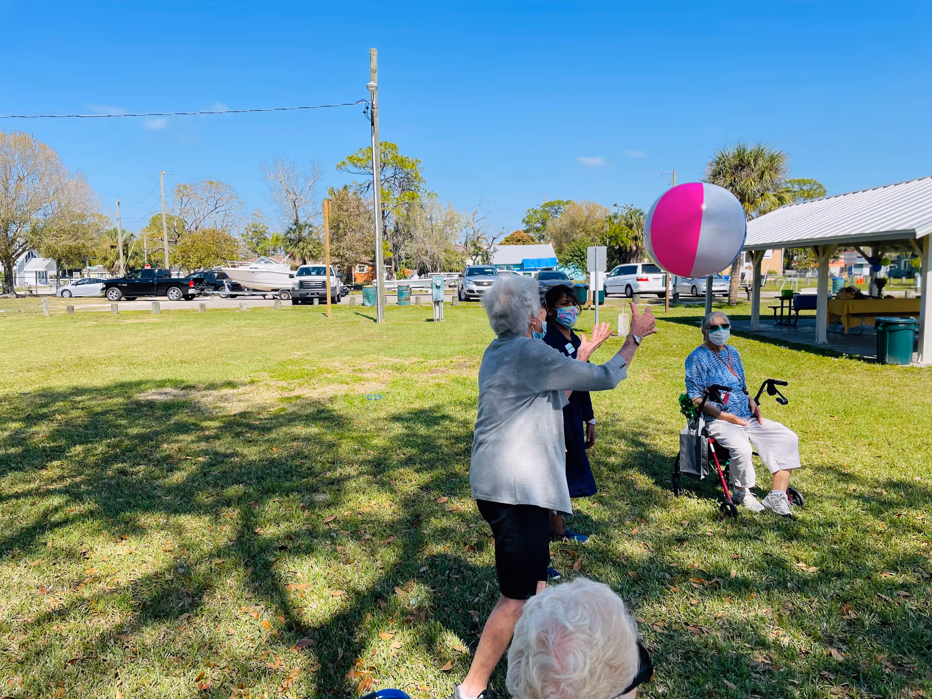 A group of elderly people outdoors on a sunny day playing with a large pink and white beach ball. One elderly woman is standing and hitting the ball, while another elderly woman is seated on a walker. A caregiver wearing a mask is standing nearby. The background shows a grassy area, parked cars, trees, and a covered pavilion.