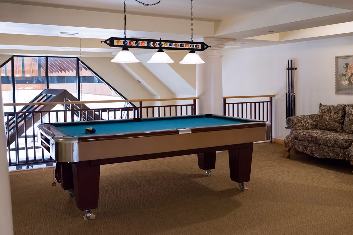 Interior room with a pool table in the center, a hanging light fixture above it with billiard balls decoration, a floral patterned couch on the right, and a rack with pool cues against the wall. Large windows and a railing are visible in the background.