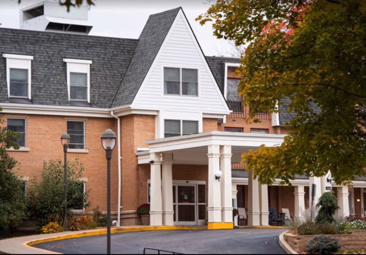 Exterior view of a senior living facility building with a covered entrance supported by white columns. The building has a brick facade with white trim and a steeply pitched roof. There are trees with autumn foliage partially framing the right side of the image.