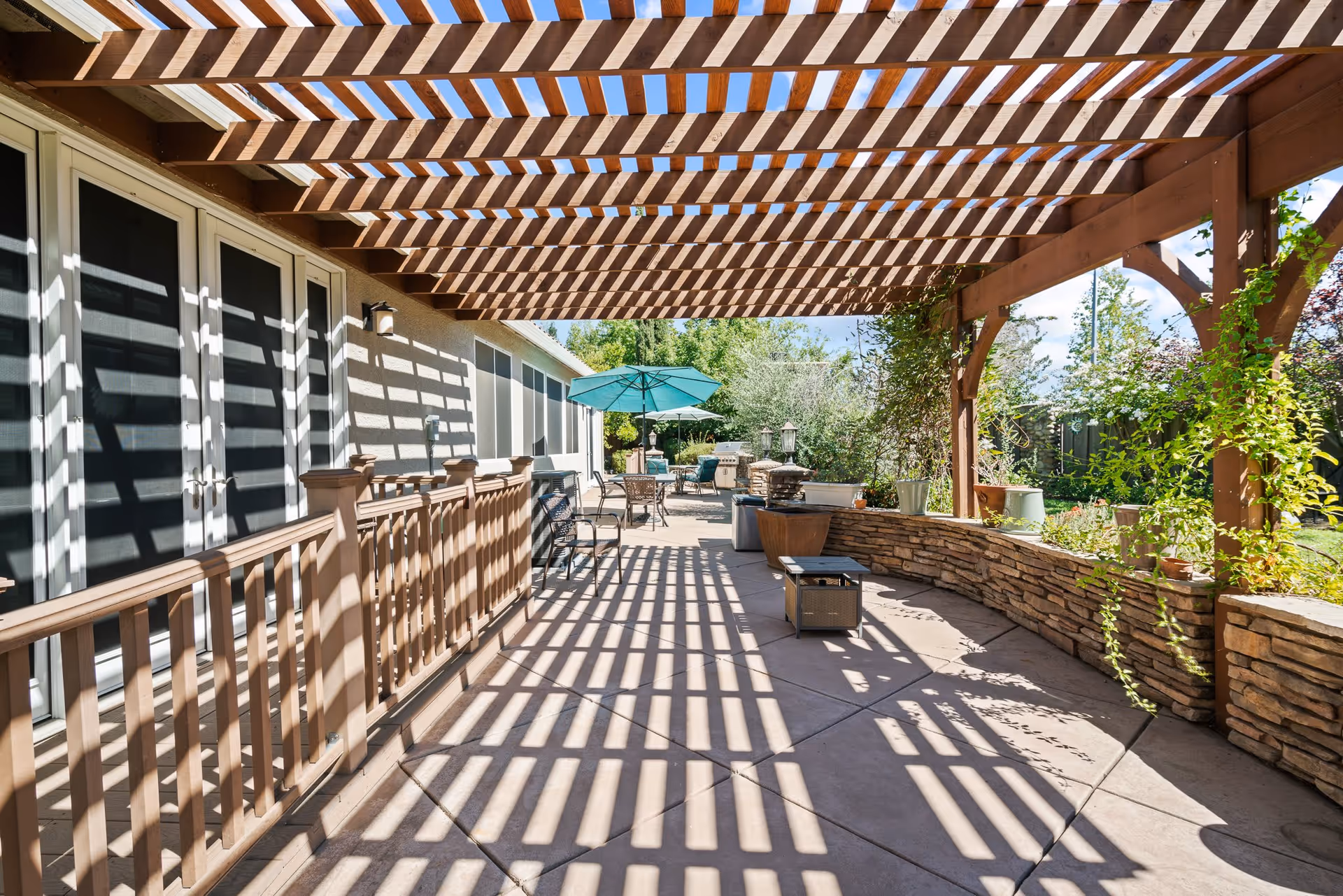 Outdoor patio area with a wooden pergola casting striped shadows on the concrete floor. There are several chairs, tables with umbrellas, potted plants, and a stone wall planter. The patio is adjacent to a building with multiple glass doors and windows.